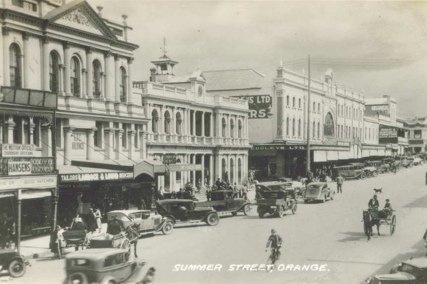 Black and white photograph of the main street of a town with horses and carts and vintage cars with two storey buildings
