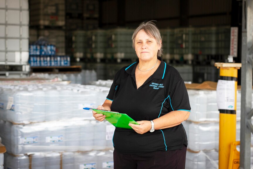 Glenda Riley holds a clipboard in front of a warehouse with supplies of donated drinking water.
