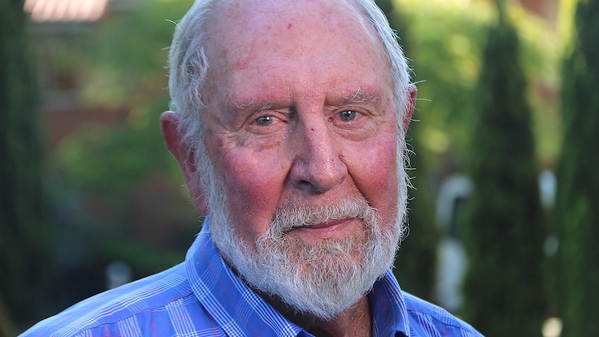 A headshot of an older man with a white beard wearing a blue checked shirt.