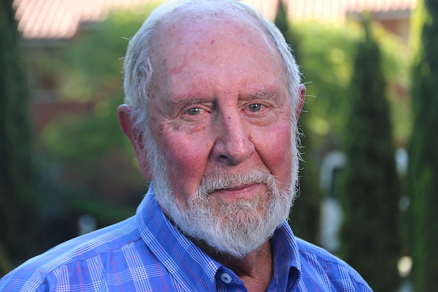 A headshot of an older man with a white beard wearing a blue checked shirt.