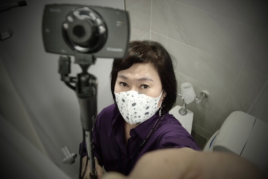 A Korean woman in a face mask holds a camera attached to a selfie stick 