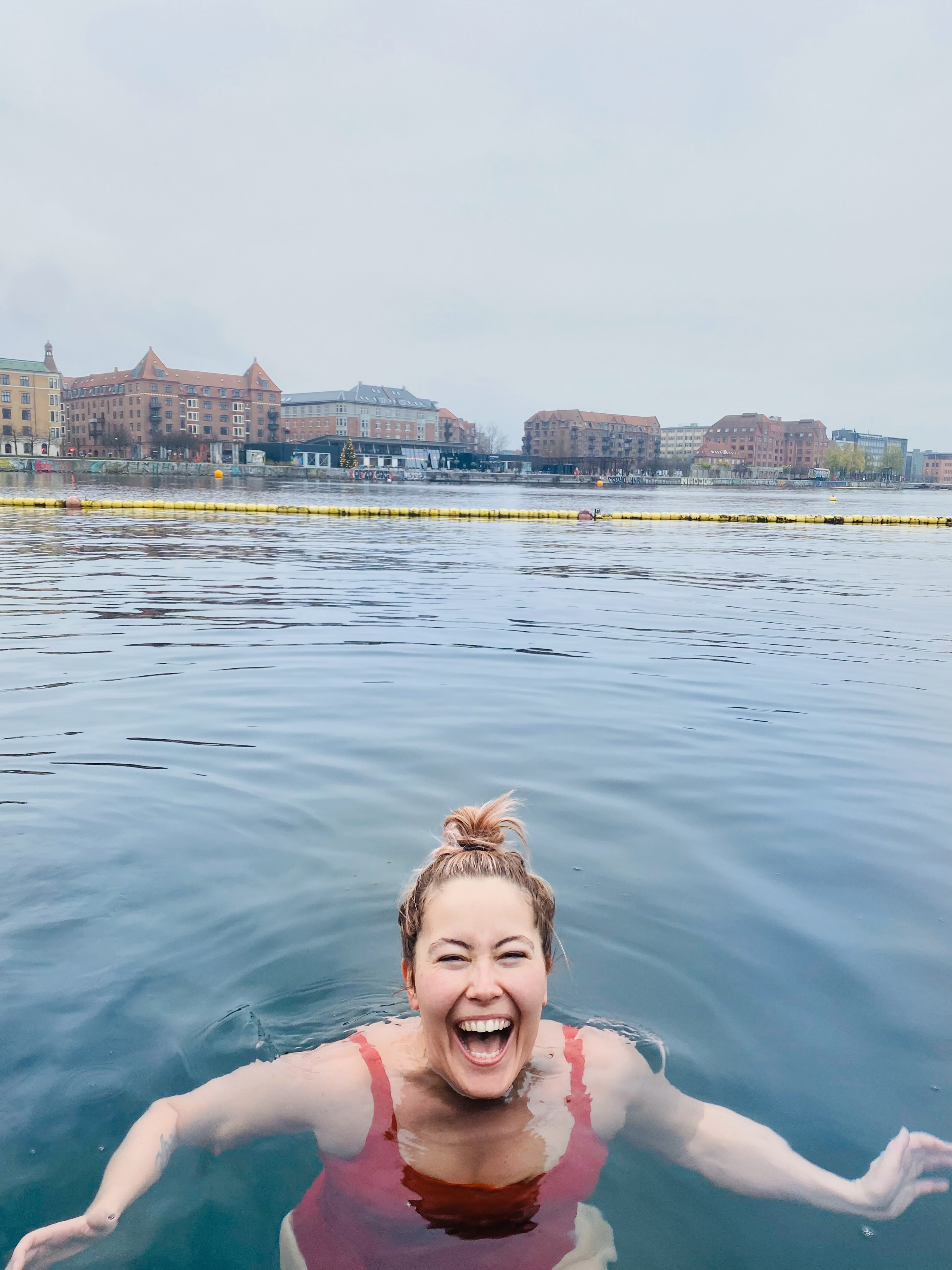 A woman in red swim suit smiles widely with just her head above water, in lake with buildings in distance behind.