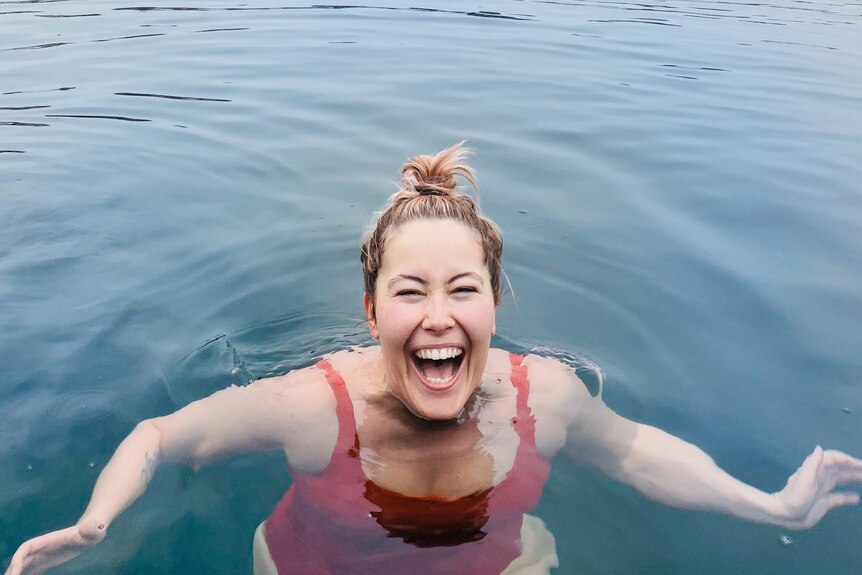 A woman in red swim suit smiles widely with just her head above water, in lake with buildings in distance behind.