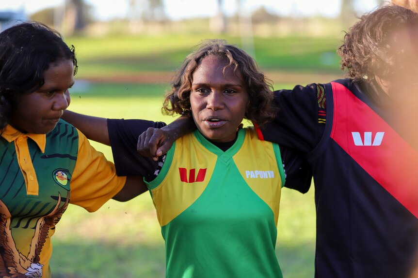 Young Aboriginal girls wearing football jumpers stand arm in arm on footbal ground