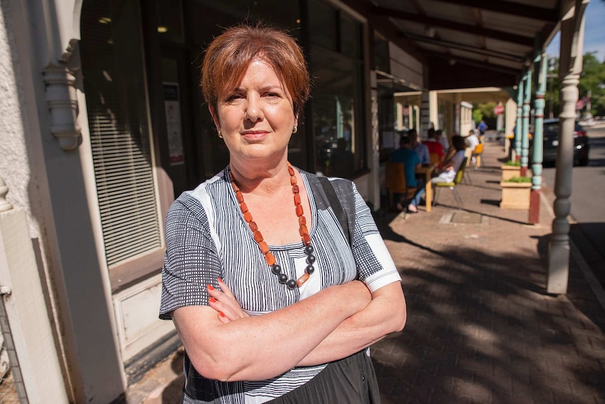 A woman stands with her arms crossed looking at the camera on a footpath.