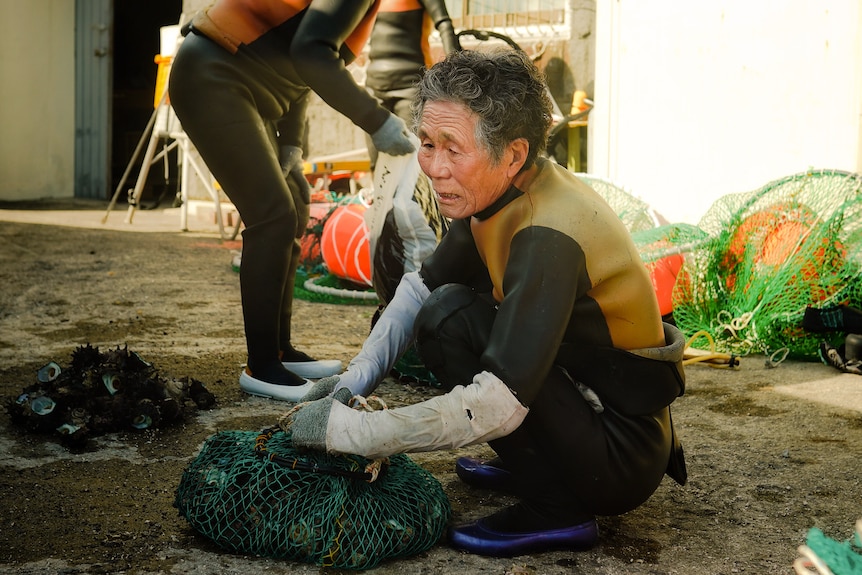 An older woman wearing diving gear holds onto a fishing net while squatting on the shore.