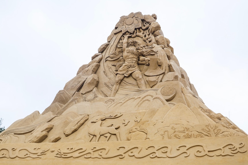 A sand sculputre shows a tall man with long beard holding his right hand up and an axe in his left hand. 