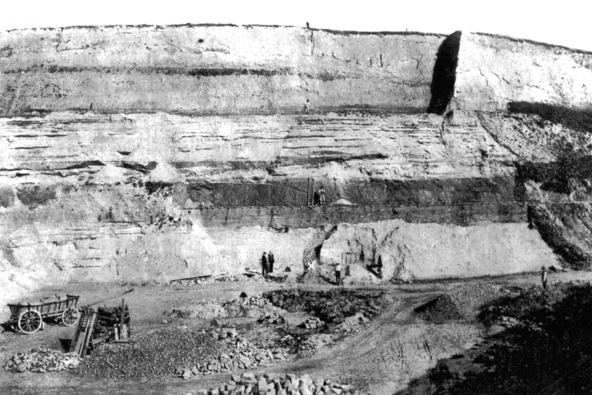 A black and while photo showing a large wall of the sand mine with a small white cross and some workers in the centre