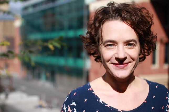 With blurred building in the background, a woman with short curly hair smiles widely with mouth closed.