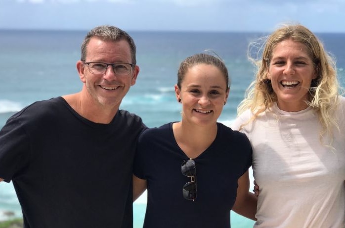 A man and three women stand in a row, with arms across each others' backs, smiling widely. Beach is in background.