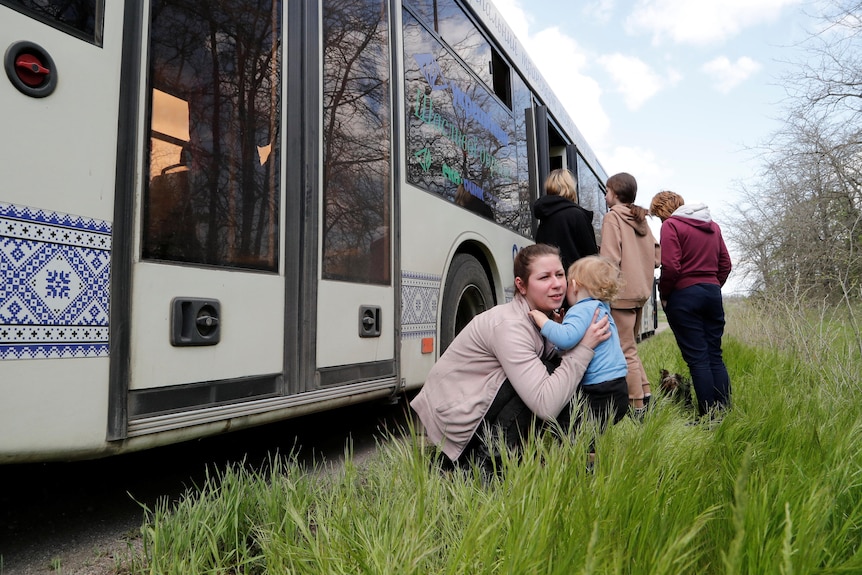 a woman crouches down next to a bus to pick up an infant