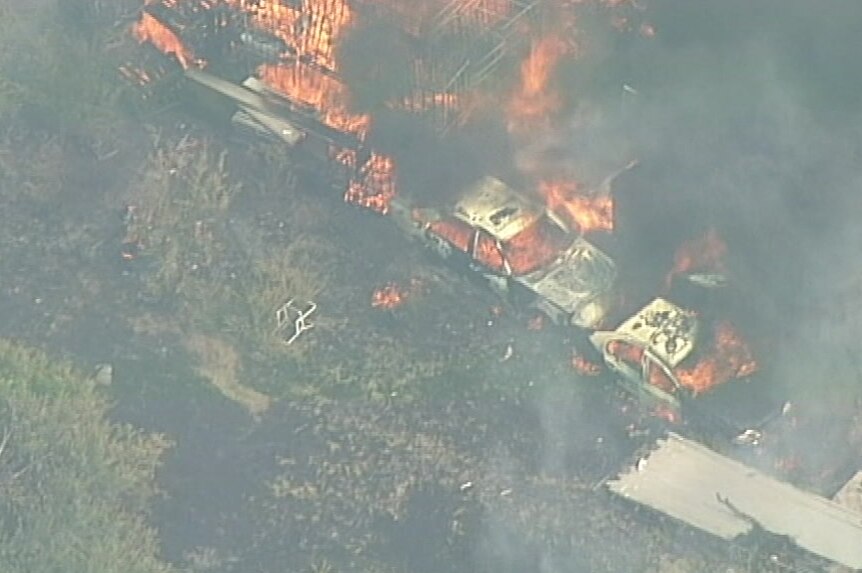 An aerial shot of two cars and structure covered in flames surrounded by grass.