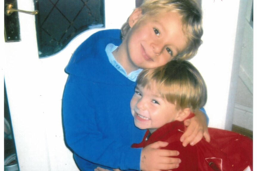 Two young blond boys hugging each other and smiling at the camera.