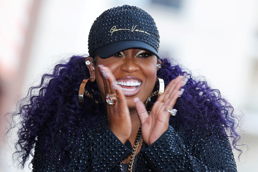 Missy Elliot clapping and smiling with curly purple hair and a cap on