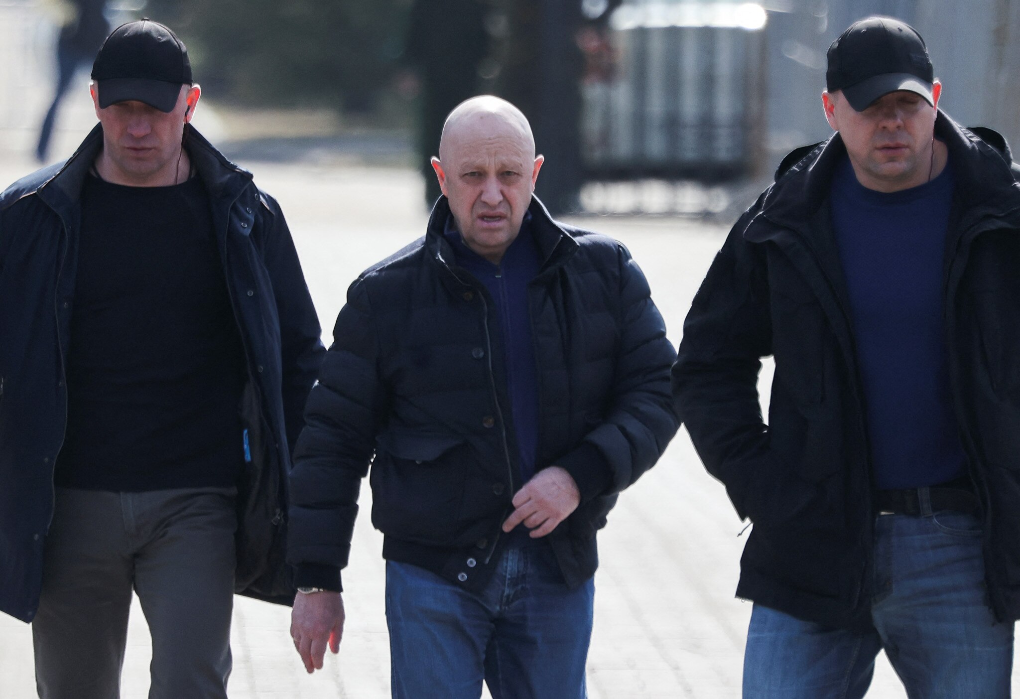 A bald man wearing a puffer jacket and jeans walks flanked by two men wearing caps and black outfits.