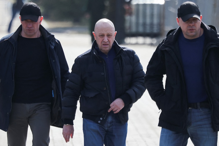 A bald man wearing a puffer jacket and jeans walks flanked by two men wearing caps and black outfits.