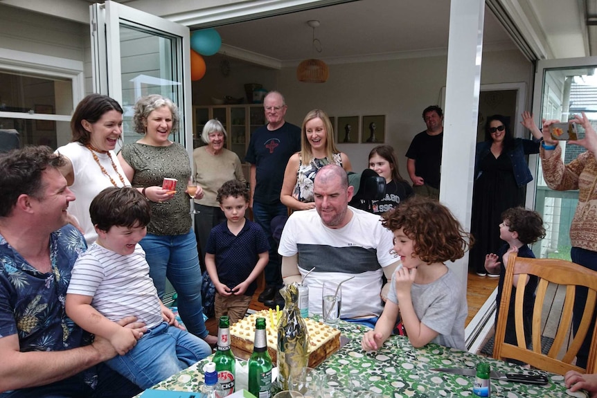 A man without limbs sits in front of a birthday cake in the backyard with a crowd of people surrounding him. They are smiling