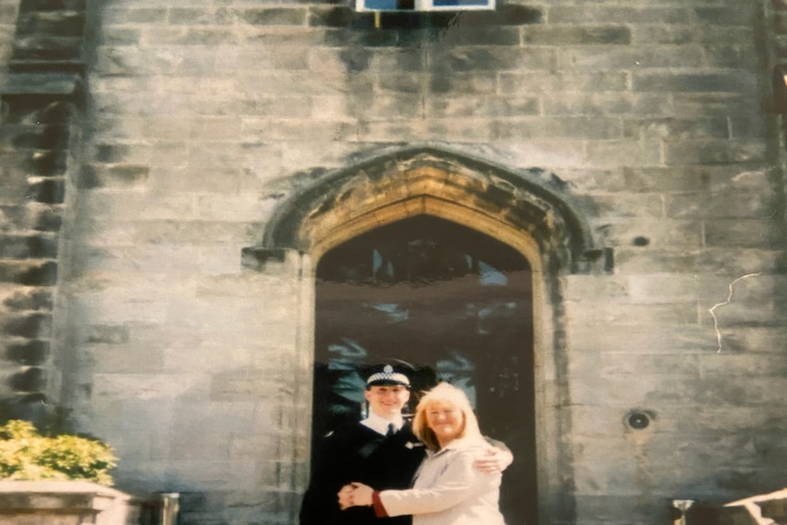Police officer in hat and uniform being hugged by a woman with fair hair outside an old building.