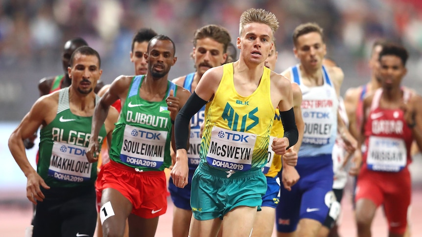 A blonde man wearing a yellow singlet runs in front of a group of men