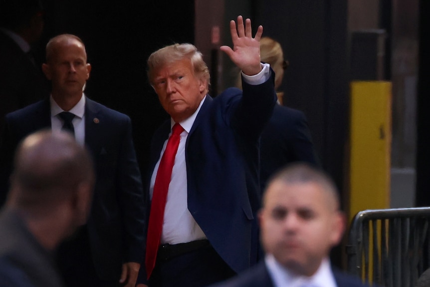 Donald Trump, wearing a blue suit and red tie, waves to a crowd as he walks past a barricade