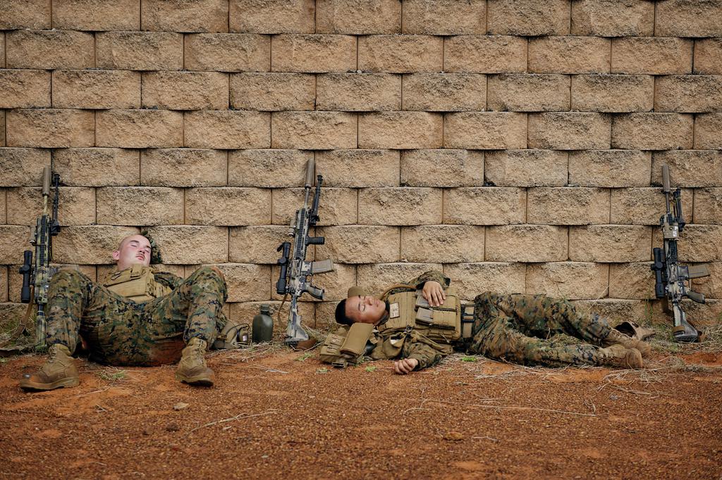Two marines in army uniform slouched sleeping against a wall, their guns propped up beside them