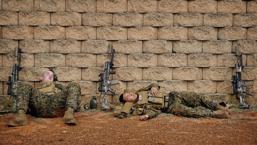 Two marines in army uniform slouched sleeping against a wall, their guns propped up beside them
