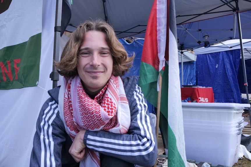 A man smiles in front fo a Palestinan flag in front of a campsite.