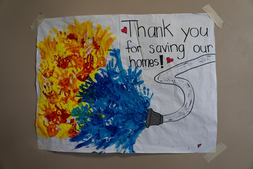 A homemade painted sign made by kids says "thank you for saving our homes" with a drawing of a fire hose.