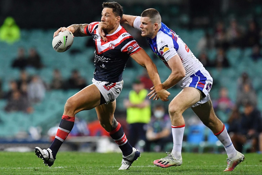 A Sydney Roosters NRL player holds the ball in his right hand as he is tackled over his right shoulder by a Newcastle opponent.