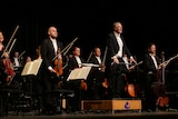A conductor and his orchestra stand up and greet a clapping audience.