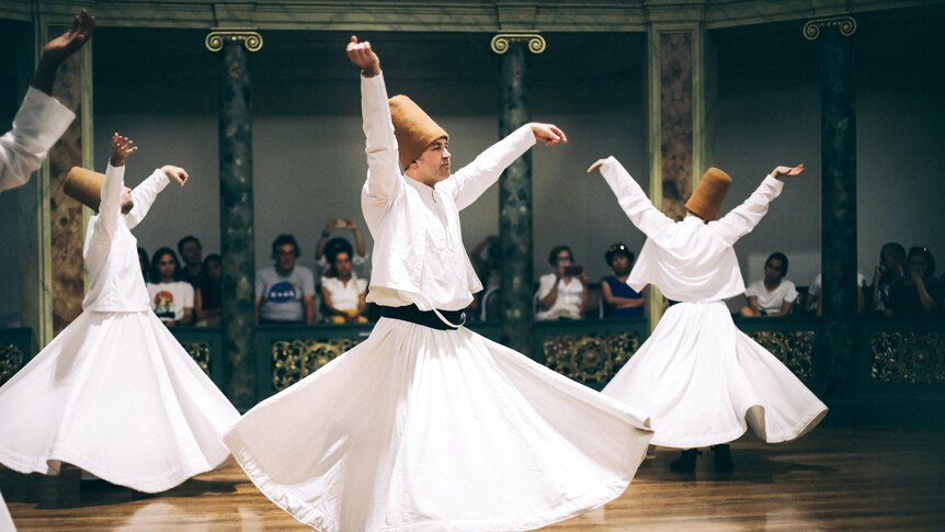 Men in white skirts wear tall brown hats and spin on the dancefloor. 