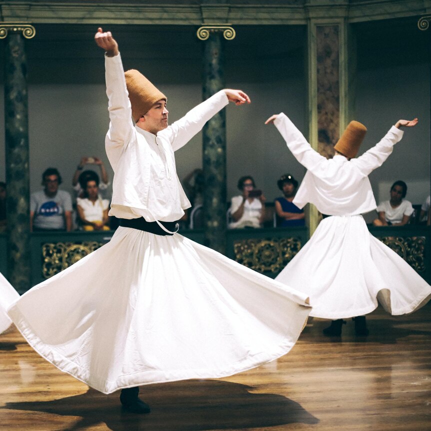 Men in white skirts wear tall brown hats and spin on the dancefloor. 