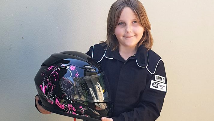 Eight-year-old Anita Board stands in a black racesuit holding her black and pink race helmet against a wall, posing for a photo.