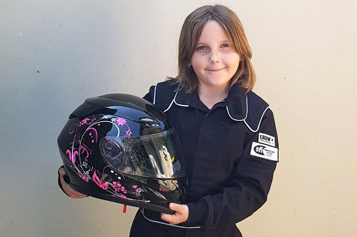 Eight-year-old Anita Board stands in a black racesuit holding her black and pink race helmet against a wall, posing for a photo.