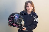 Eight-year-old Anita Board stands in a black racesuit holding her black and pink race helmet against a wall, posing for a photo.