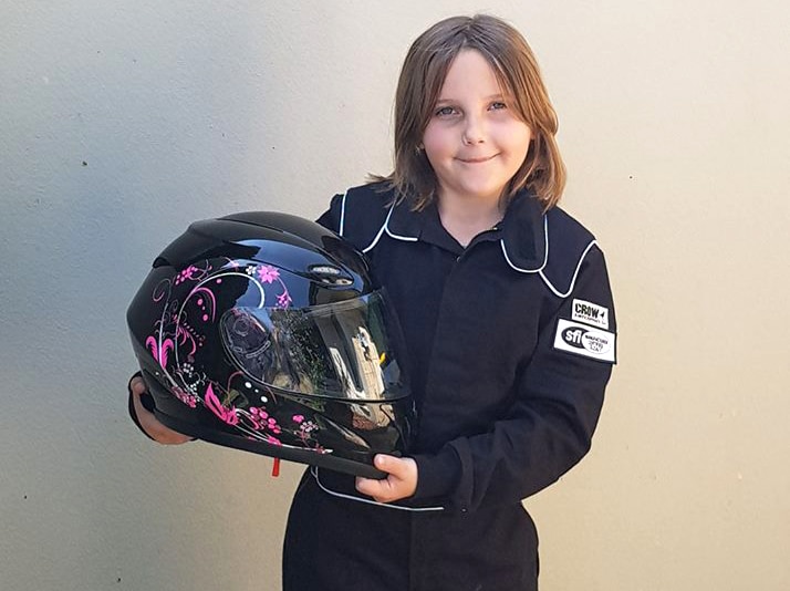 Eight-year-old Anita Board stands in a black racesuit holding her black and pink race helmet against a wall, posing for a photo.