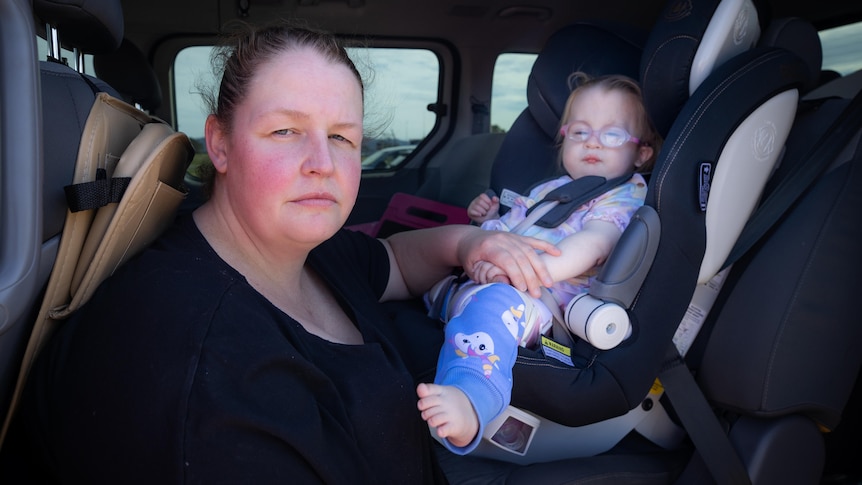 A mother holds the hand of her toddler, who is in a child car restraint. 