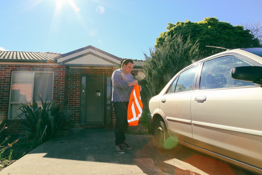 A man is pictured in front of his house, holding a high-vis vest, standing beside his car.