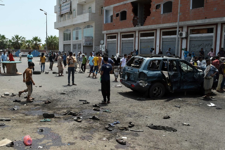 People stand at the site of a bombing near a damaged car.