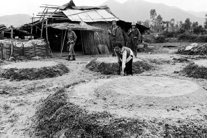 A woman at a circular gravesite mound with soldiers standing behind her.