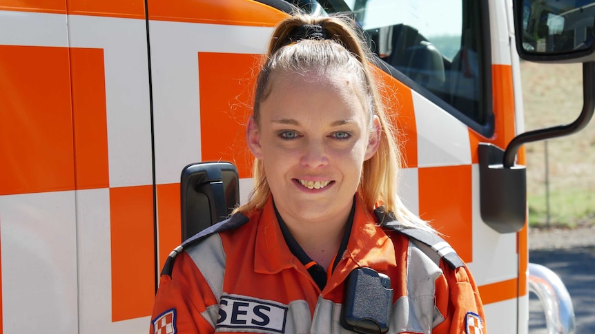 A smiling women with a high blonde ponytail in an orange jumpsuit stands in front of an emergency service truck.