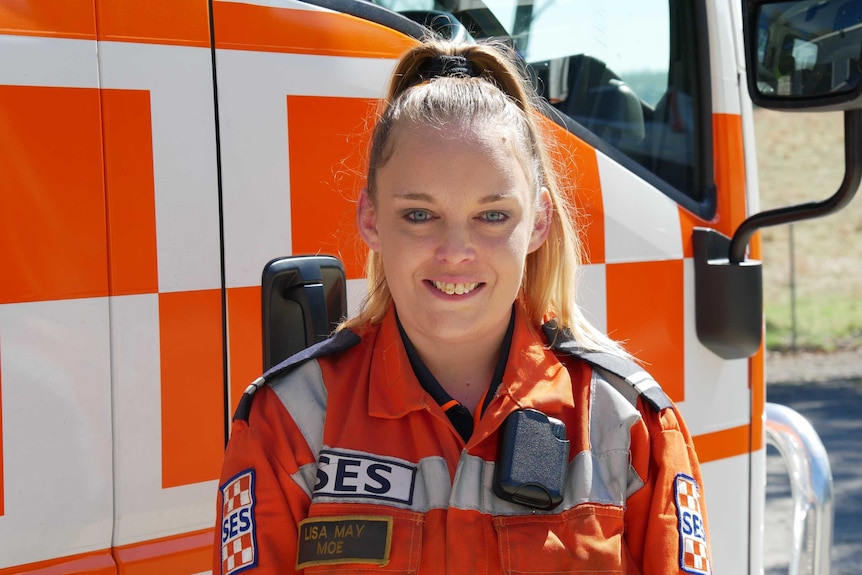 A smiling women with a high blonde ponytail in an orange jumpsuit stands in front of an emergency service truck.