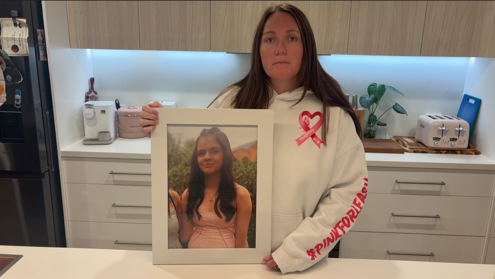 A woman holds a framed photo of Annalyse Blyton. She is wearing a jumper with a pink heart logo and Pink for Leasy written on it