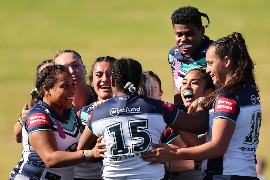 A Cowboys NRLW player with her back to camera is surrounded by smiling teammates after her try.
