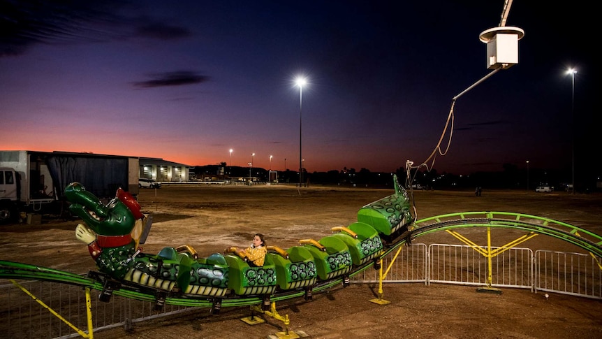 Against a navy and pink sky, a green rollercoaster carries one young person around a track.