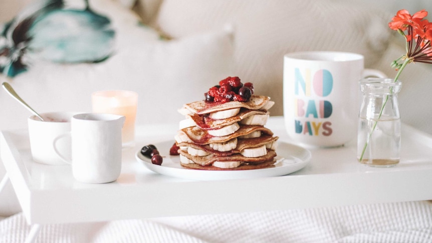 A stack of pancakes with banana and berries sits on a white bedspread.