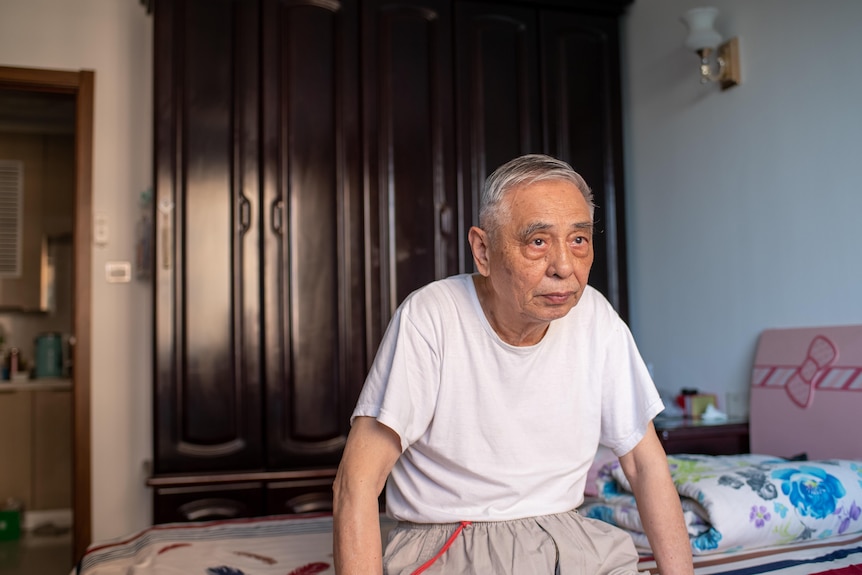 An old man sits on a bed staring off into the distance.