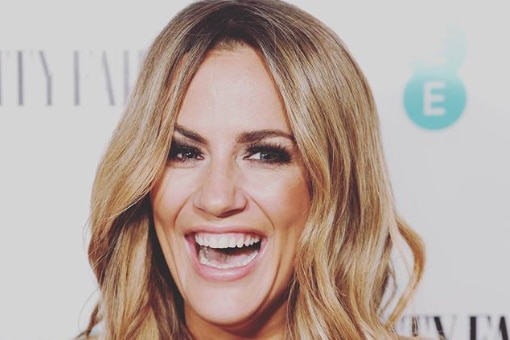 Caroline Flack smiles with her hair curled in front of a Vanity Fair board.
