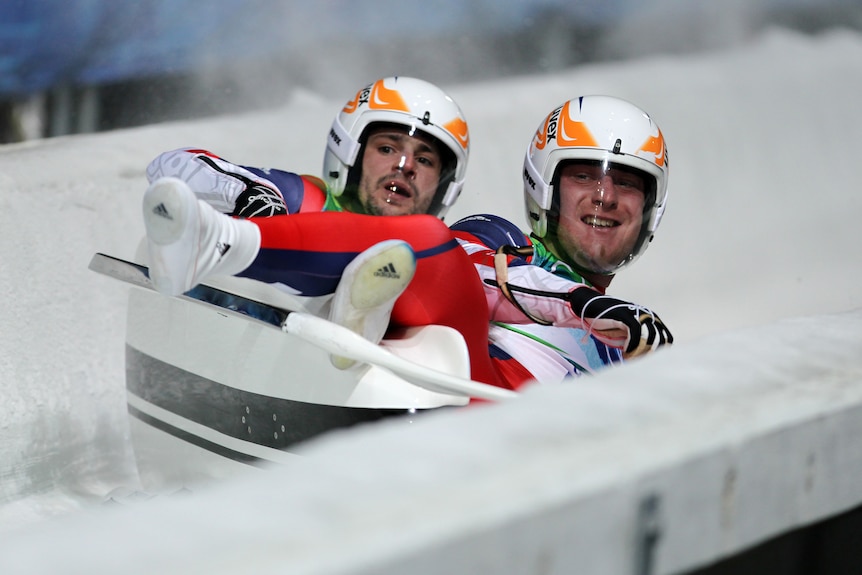 Two luge competitors look over the side of their sled with pained expressions on their face