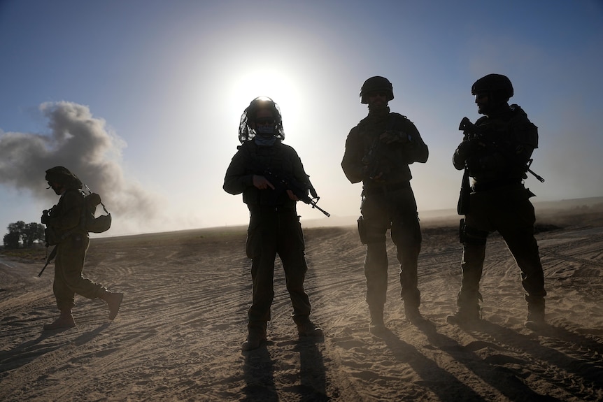 The silhouettes of four soldiers holding guns in a desert, with the sun in the background
